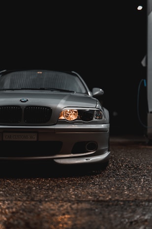 A sleek silver car is prominently displayed, with its headlights on, casting a warm glow against the dark background. The car's front grille and emblem are visible, along with detailed reflections on the smooth surface of the car body. The environment is dimly lit, emphasizing the polished look of the vehicle.