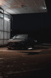 A car is parked in a dimly lit, covered area at night. Overhead lighting casts a soft glow on the vehicle, creating reflections on its surface. The environment appears to be a car wash or service area with metallic siding and a wet ground.