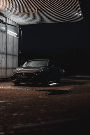 A car is parked in a dimly lit, covered area at night. Overhead lighting casts a soft glow on the vehicle, creating reflections on its surface. The environment appears to be a car wash or service area with metallic siding and a wet ground.
