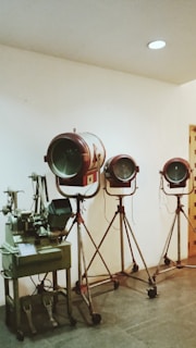 Three vintage film spotlights on tripod stands are arranged in a row against a plain white wall. To the left, there is an old-fashioned film editing machine.