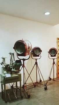 Three vintage film spotlights on tripod stands are arranged in a row against a plain white wall. To the left, there is an old-fashioned film editing machine.