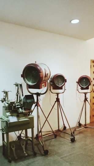 Three vintage film spotlights on tripod stands are arranged in a row against a plain white wall. To the left, there is an old-fashioned film editing machine.