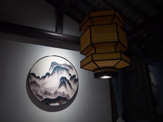 A traditional Asian lantern with a geometric design hangs indoors. Next to it is a circular painting depicting stylized mountains and forests, reminiscent of classic Asian art. The wall is painted white, and the setting appears calm and serene.