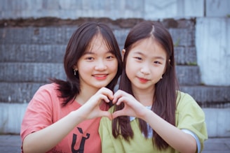 Two young people are seated in front of a staircase, facing the camera and smiling warmly. They are forming a heart shape with their hands together, symbolizing affection or friendship. Both are casually dressed, with one wearing a pink top and the other in a light green shirt.