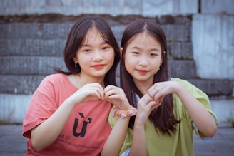 Two young individuals sitting together on steps, forming heart shapes with their hands. One wears a red shirt, and the other wears a light green top. They both have long hair and are looking at the camera, appearing relaxed and content.