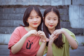 Two young individuals sitting together on steps, forming heart shapes with their hands. One wears a red shirt, and the other wears a light green top. They both have long hair and are looking at the camera, appearing relaxed and content.