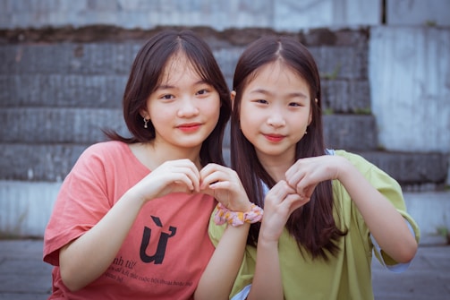 Two young individuals sitting together on steps, forming heart shapes with their hands. One wears a red shirt, and the other wears a light green top. They both have long hair and are looking at the camera, appearing relaxed and content.