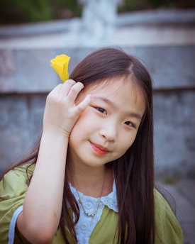 A young girl with long, dark hair is smiling softly and holding a small yellow flower next to her face. She is wearing a green shirt with a white collar and a necklace, against a blurred background.