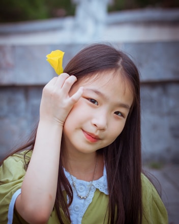 A young girl with long, dark hair is smiling softly and holding a small yellow flower next to her face. She is wearing a green shirt with a white collar and a necklace, against a blurred background.