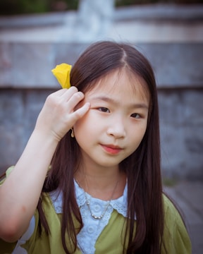 A young girl with long dark hair holds a yellow flower near her temple. She is wearing a green top with a white collar and a necklace with small pearls.