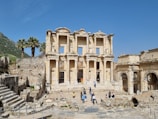 A well-preserved ancient building facade with multiple columns and intricate stone carvings dominates the scene. In front, a group of tourists, some walking and others standing, are exploring the historic site. Palm trees and stone ruins are visible in the background against a clear blue sky.