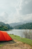 A vibrant orange tent pitched beside a clear mountain lake under a bright blue sky.