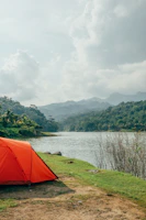 Drone shot of a vibrant orange tent set up beside a turquoise river surrounded by lush forest.