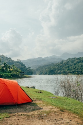 A vibrant orange tent pitched beside a clear mountain lake under a bright blue sky.