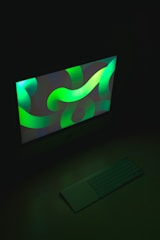 A desktop computer screen featuring a vibrant digital artwork with swirling green and blue shapes, casting a colorful glow. In front of the monitor sits a sleek, minimalistic keyboard and touchpad, subtly illuminated by the screen's light. The overall setting is dark, highlighting the luminous display.