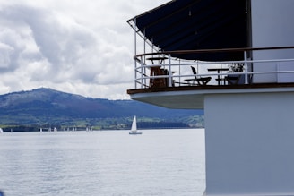 A serene lakeside scene featuring a partially visible modern building with a patio, containing chairs and a table. In the background, a picturesque landscape includes a calm lake with sailboats scattered across the water, and a hilly terrain under a cloudy sky.