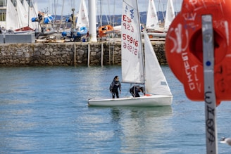 Two people are sailing a small white sailboat in a calm bay. The background is lined with a dock that has several moored boats and yachts. There is a prominent orange lifebuoy with text on it in the foreground to the right. The water reflects the light, creating a serene and peaceful scene.