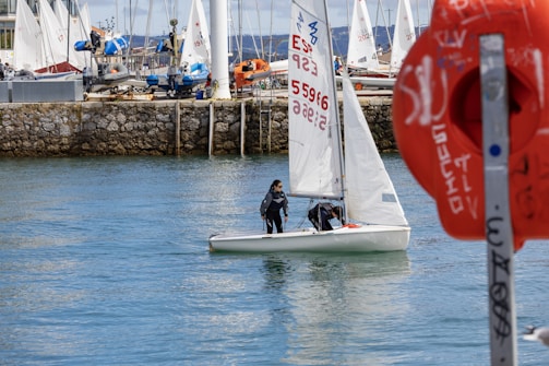 Two people are sailing a small white sailboat in a calm bay. The background is lined with a dock that has several moored boats and yachts. There is a prominent orange lifebuoy with text on it in the foreground to the right. The water reflects the light, creating a serene and peaceful scene.