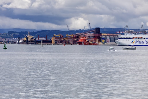 A coastal industrial area is visible with several ships and boats, including a large ferry and smaller vessels. Behind these, there are buildings and structures, possibly for loading and unloading cargo, with cranes and industrial equipment. The sky is overcast with patches of clouds, and hilly terrain can be seen in the background.