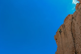 A person wearing a bright green shirt is rock climbing on a large, rugged cliff under a clear blue sky. The climber is secured with ropes and appears to be ascending a steep vertical surface composed of reddish-brown rock.