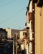 A narrow urban street with a mix of modern and older buildings lines the frame. A series of streetlights and utility wires stretch above, leading toward a large building. The Ukrainian flag is visible on a distant building, while several vehicles and a pedestrian sign indicate active city life.