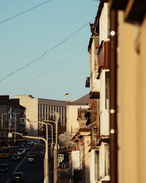 A narrow urban street with a mix of modern and older buildings lines the frame. A series of streetlights and utility wires stretch above, leading toward a large building. The Ukrainian flag is visible on a distant building, while several vehicles and a pedestrian sign indicate active city life.