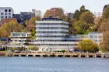 A modern, multi-story building labeled GEOMAR located by a waterfront. The building is surrounded by trees and adjacent structures. The scene includes a dock or walkway along the water with people walking and relaxing.