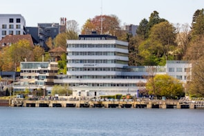A modern, multi-story building labeled GEOMAR located by a waterfront. The building is surrounded by trees and adjacent structures. The scene includes a dock or walkway along the water with people walking and relaxing.
