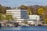 A multi-story white office building is situated by the water, with several satellite dishes on the roof. Surrounding the building are numerous trees, some with fresh green foliage, hinting at a springtime setting. In the foreground, a marina with several small boats and a dock can be seen, alongside a group of people walking.