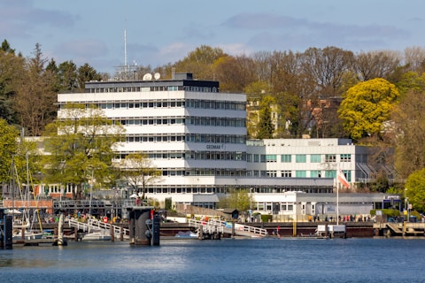 A multi-story white office building is situated by the water, with several satellite dishes on the roof. Surrounding the building are numerous trees, some with fresh green foliage, hinting at a springtime setting. In the foreground, a marina with several small boats and a dock can be seen, alongside a group of people walking.