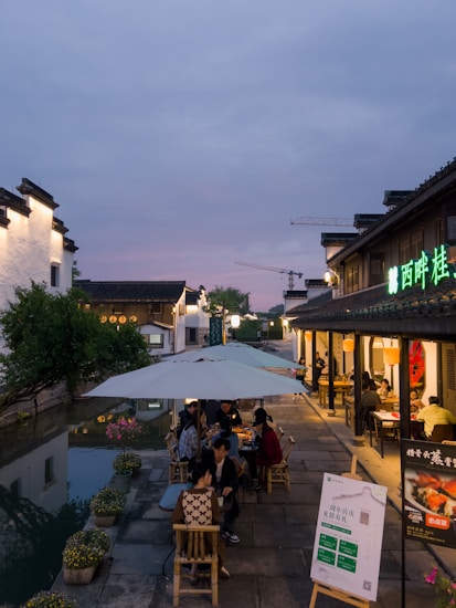 An outdoor dining area set alongside a historic-style building with Chinese architectural elements is visible. Several people are seated under large umbrellas, engaging in conversation. The path is flanked by a serene waterway lined with greenery and flowers. A colorful neon sign in Chinese characters adorns one building. Soft evening lighting creates a calm atmosphere, and cranes are visible in the distant skyline, hinting at ongoing development.
