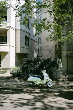 A pale green scooter is parked on a shaded street in front of a modern building with light-colored stone walls. The building has arched windows and is partially obscured by leafy trees. There is dense greenery, including ivy-covered fences, creating a peaceful, urban atmosphere.