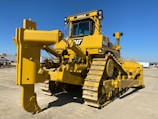 A large yellow bulldozer with a powerful front blade is parked in a construction area under a clear blue sky. The vehicle's tracks and intricate mechanical components are visible, emphasizing its robust construction. In the background, there are other trucks and construction equipment.