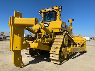 A large yellow bulldozer with a powerful front blade is parked in a construction area under a clear blue sky. The vehicle's tracks and intricate mechanical components are visible, emphasizing its robust construction. In the background, there are other trucks and construction equipment.