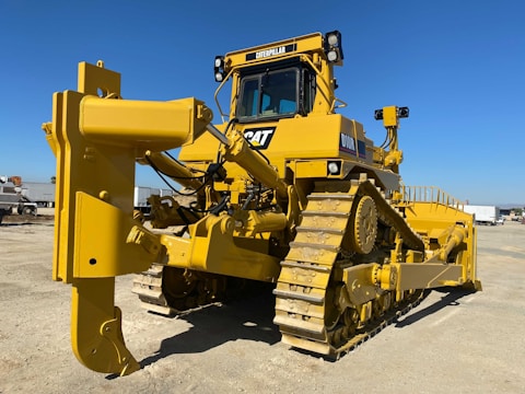 A large yellow bulldozer with a powerful front blade is parked in a construction area under a clear blue sky. The vehicle's tracks and intricate mechanical components are visible, emphasizing its robust construction. In the background, there are other trucks and construction equipment.