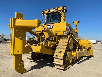 A large yellow bulldozer with a powerful front blade is parked in a construction area under a clear blue sky. The vehicle's tracks and intricate mechanical components are visible, emphasizing its robust construction. In the background, there are other trucks and construction equipment.
