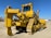 A large yellow bulldozer with a powerful front blade is parked in a construction area under a clear blue sky. The vehicle's tracks and intricate mechanical components are visible, emphasizing its robust construction. In the background, there are other trucks and construction equipment.