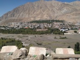 A town is nestled against a barren mountain backdrop, with a river in the foreground. The town features a mix of colorful buildings surrounded by green trees. The area appears dry with rugged and rocky terrain.