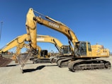 A row of large, yellow excavators with the brand 'Deere' prominently displayed are parked on a gravel lot. These heavy machinery vehicles have bucket attachments and are positioned under a clear blue sky.