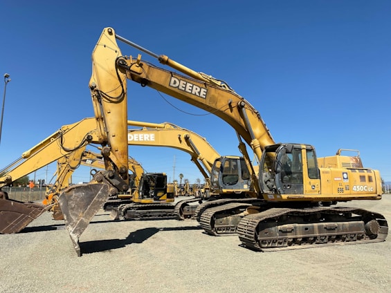 A row of large, yellow excavators with the brand 'Deere' prominently displayed are parked on a gravel lot. These heavy machinery vehicles have bucket attachments and are positioned under a clear blue sky.