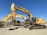 A row of large, yellow excavators with the brand 'Deere' prominently displayed are parked on a gravel lot. These heavy machinery vehicles have bucket attachments and are positioned under a clear blue sky.