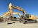 A row of large, yellow excavators with the brand 'Deere' prominently displayed are parked on a gravel lot. These heavy machinery vehicles have bucket attachments and are positioned under a clear blue sky.