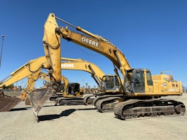 A row of large, yellow excavators with the brand 'Deere' prominently displayed are parked on a gravel lot. These heavy machinery vehicles have bucket attachments and are positioned under a clear blue sky.