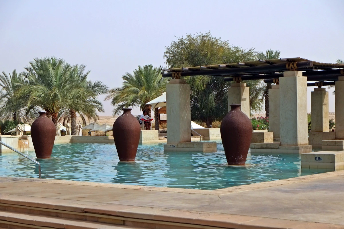 Three tall clay urns standing in a turquoise pool beneath date palms and a shaded pergola
