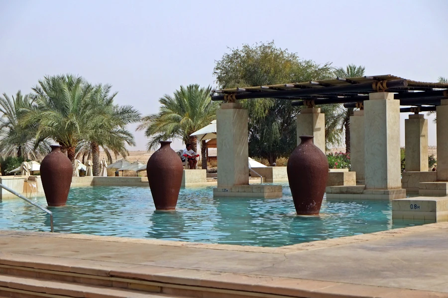 Three tall clay urns standing in a turquoise pool beneath date palms and a shaded pergola