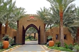 An entrance gate with tall, arched wooden doors flanked by palm trees on both sides. The gate is part of an adobe-style structure with a logo and text that reads 'BAB AL SHAMS' above the entrance. In front of the gates, there are large terracotta pots with plants. The atmosphere appears tranquil with lush greenery beyond the entrance.