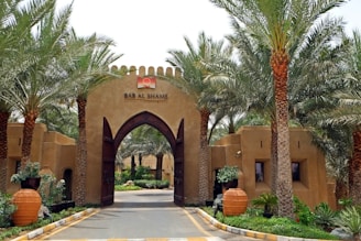 An entrance gate with tall, arched wooden doors flanked by palm trees on both sides. The gate is part of an adobe-style structure with a logo and text that reads 'BAB AL SHAMS' above the entrance. In front of the gates, there are large terracotta pots with plants. The atmosphere appears tranquil with lush greenery beyond the entrance.