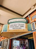 A colorful entrance sign to Neptune's Kingdom displays various attractions including mini golf, arcade, magic shop, pool, and restrooms. Surrounding the sign are decorative elements in teal and gold, with additional vibrant architectural features visible.