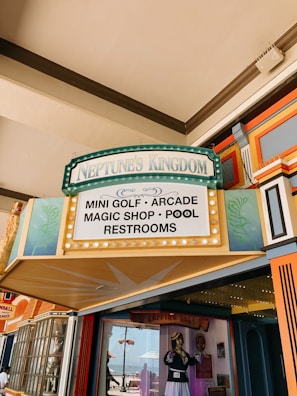 A colorful entrance sign to Neptune's Kingdom displays various attractions including mini golf, arcade, magic shop, pool, and restrooms. Surrounding the sign are decorative elements in teal and gold, with additional vibrant architectural features visible.