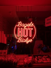 A neon sign glows prominently with the words 'Bagels HOT Bialys' in bright red. Below the sign, various packaged baked goods are displayed on a counter. The interior of the establishment is dimly lit, giving a cozy and atmospheric vibe.